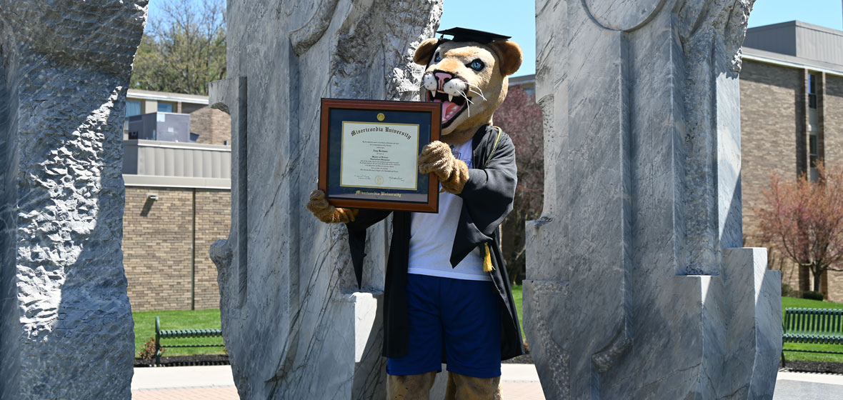 school mascot dressed in a graduation cap and gound holding a diploma