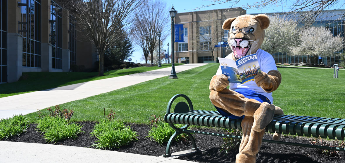 mascot sitting on a bench outside on campus, reading